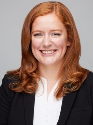 A smiling woman with red hair, wearing a black blazer and white blouse, against a neutral background.
